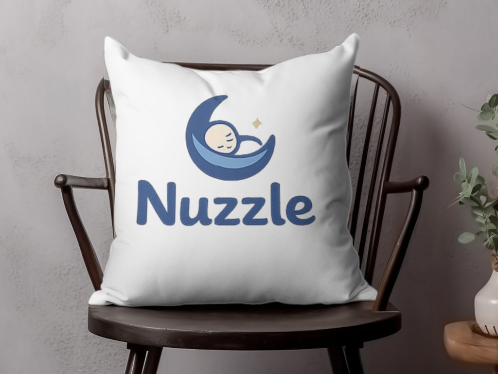 White pillow with blue "Nuzzle" text and a sleeping baby on a crescent moon logo.
